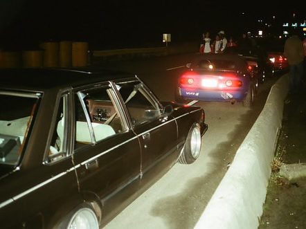 Nighttime street racing scene with parked cars and people socializing on the roadside under streetlights.