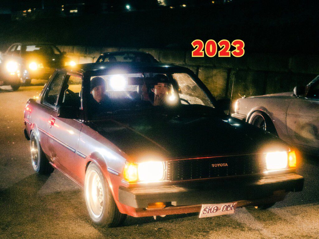 Vintage Toyota at night, illuminated headlights, captures nostalgic car culture, 2023 atmosphere.