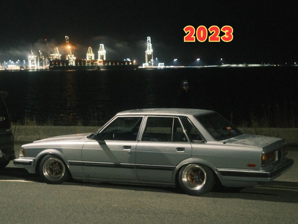 Vintage silver sedan parked at night by a waterfront with illuminated cranes in the background, marked 2023.