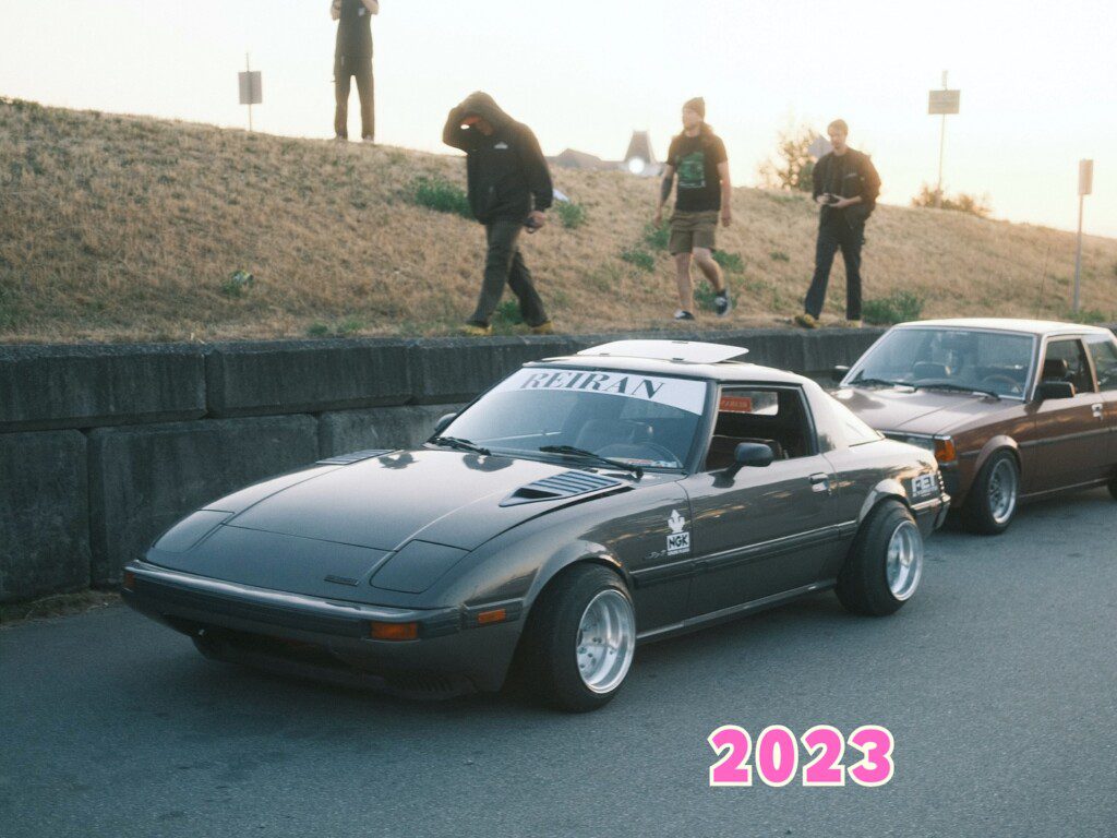 Retro car meetup 2023: Classic gray sports car parked on street with onlookers in the background.