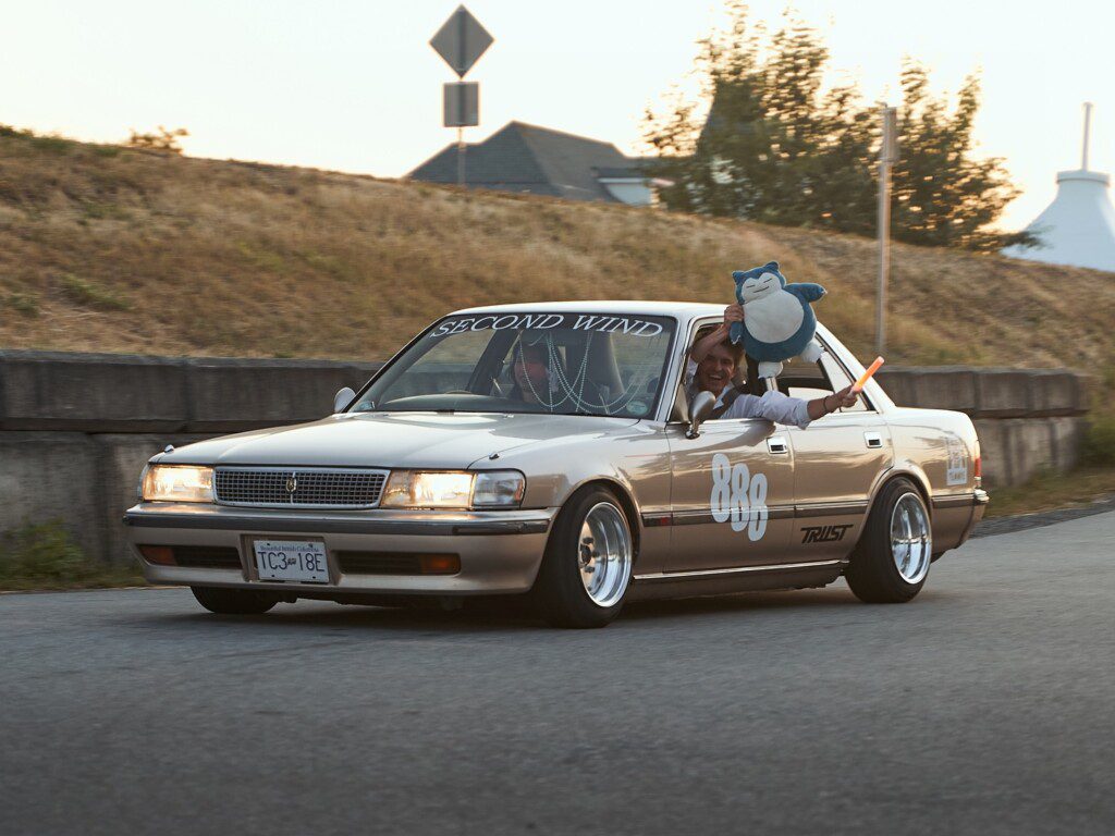 Vintage car driving on road with person holding plush toy and waving stick through window.