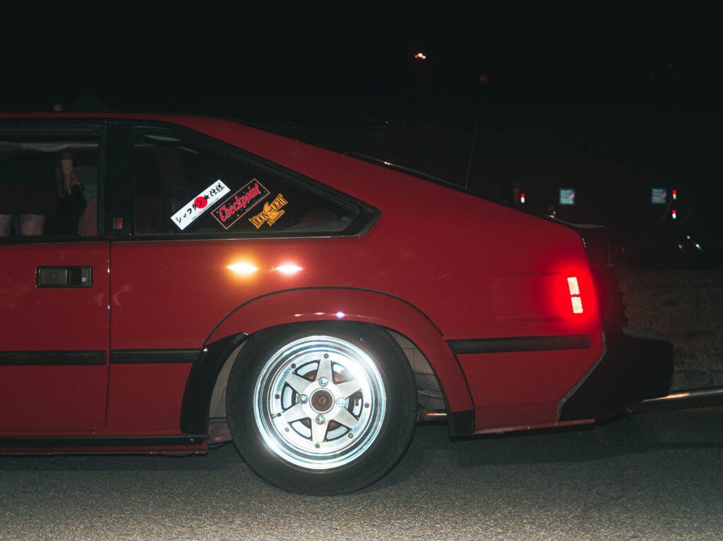Red vintage car with chrome wheels at night, featuring rear window decals.