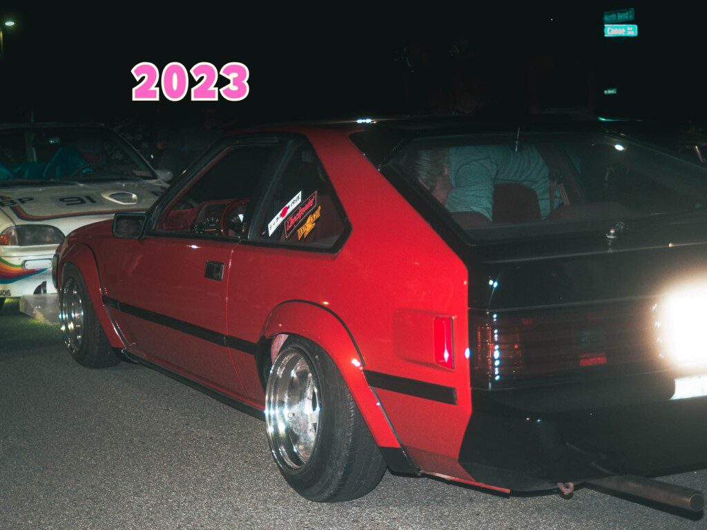 Red sports car at a night car meetup, 2023 text overlay, showcasing sleek design and retro vibe.