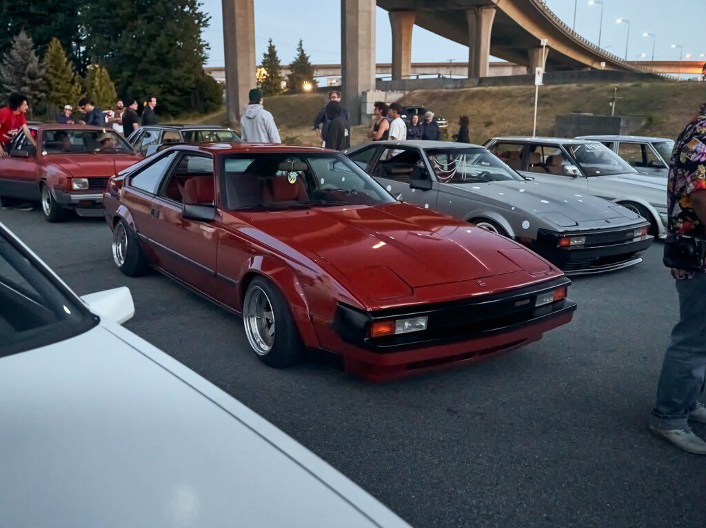 Red vintage sports car at a car meet, surrounded by classic cars and enthusiasts under a highway bridge.