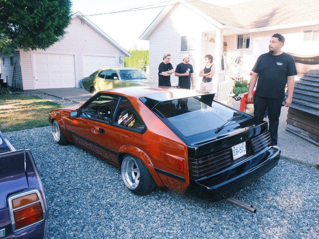 Red vintage sports car parked on gravel, surrounded by people chatting, with a garage and house in the background.