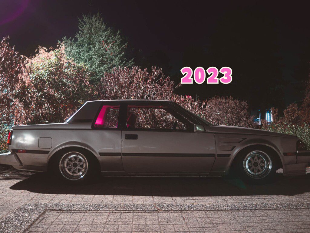 Vintage car parked at night with 2023 text overlay, surrounded by illuminated foliage.