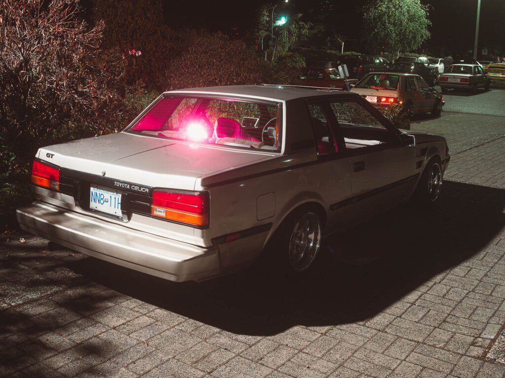 Vintage Toyota Celica parked at night under streetlights. Retro car with lit taillights, quiet urban setting.