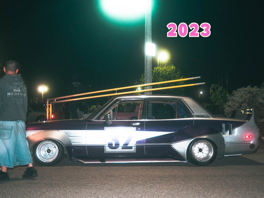 Classic customized car at night with a racing number, 2023 graffiti. Street lights and person in background.