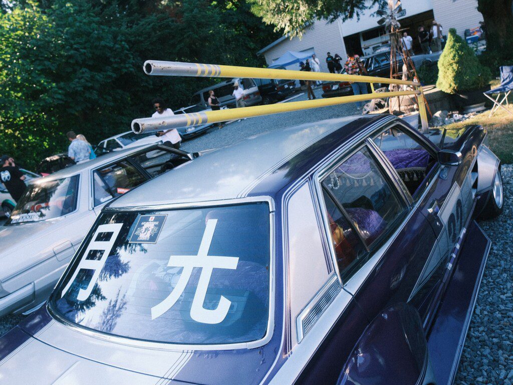 Customized car with Japanese text on window and yellow pipes at a car meet surrounded by enthusiasts.