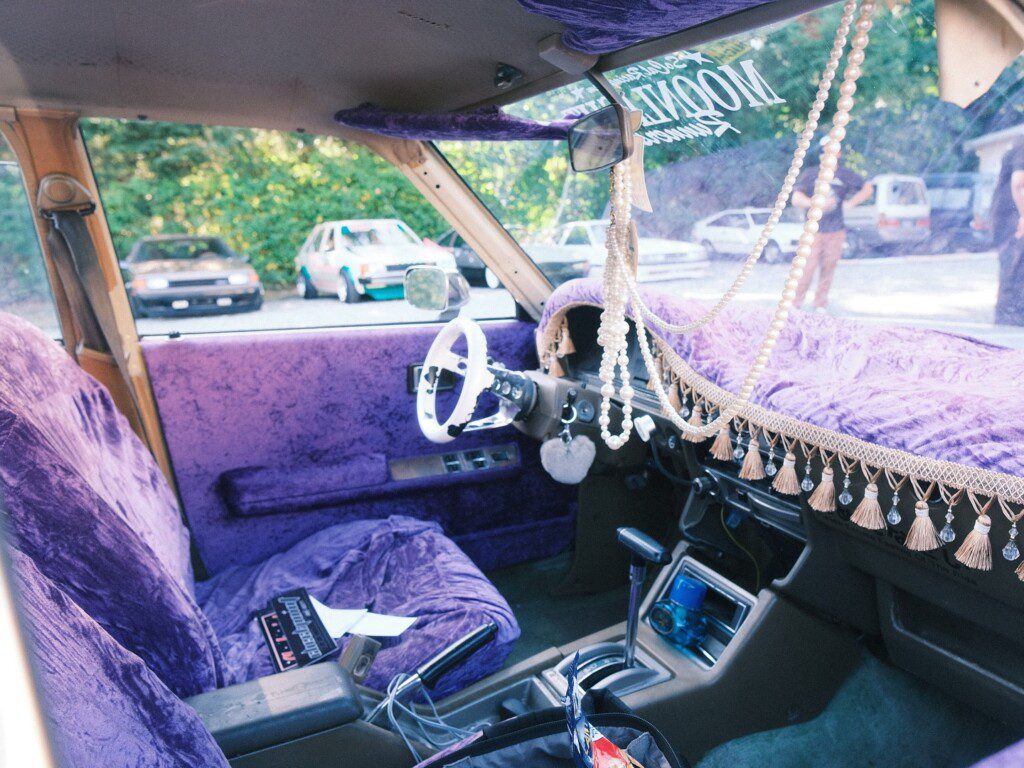 Vintage car interior with purple velvet, tassels, and beads on dashboard, parked outdoors with trees in the background.