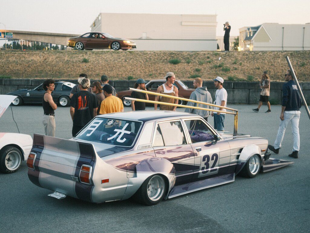 Custom vintage car at a gathering with people interacting in the background, showcasing Japanese street racing culture.