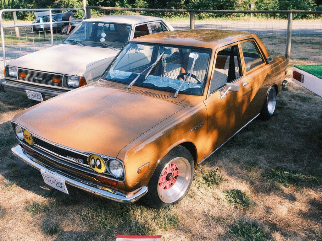 Vintage Datsun car with round headlights and a brown exterior parked beside an off-white vehicle at a car show.