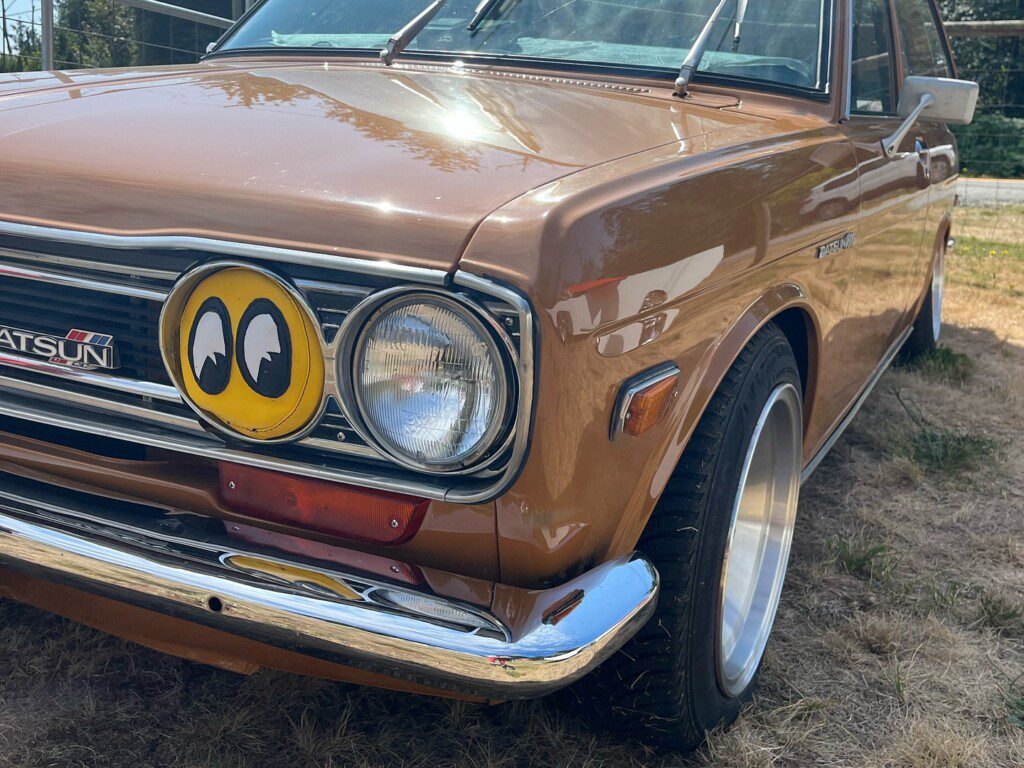 Vintage brown Datsun 510 with a custom smiley face headlight cover, parked on grass in sunlight.
