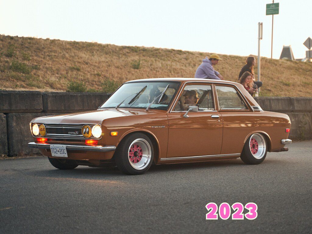 Vintage brown Datsun car driving on road, 2023, people sitting on wall in background. Classic vehicle enthusiasts gathering.