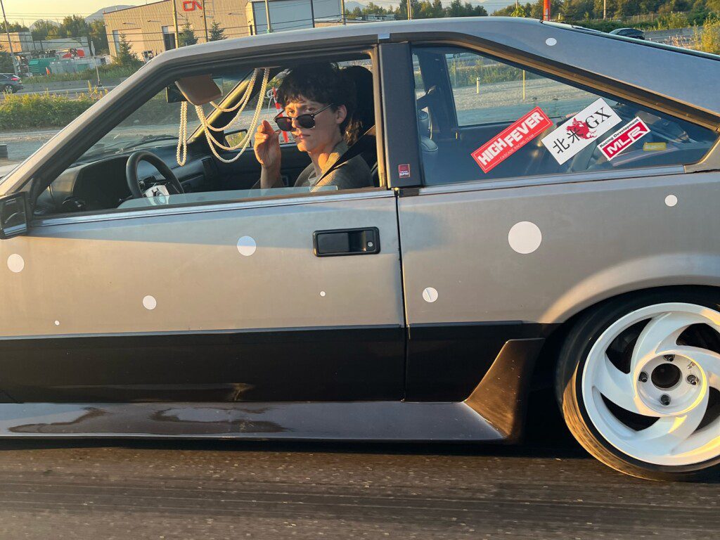Retro car cruising, driver wearing sunglasses, stylish decals on car window, scenic background during sunset drive.
