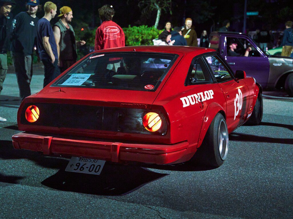 Red sports car with Dunlop branding and number 130 at a nighttime car meetup, surrounded by people.