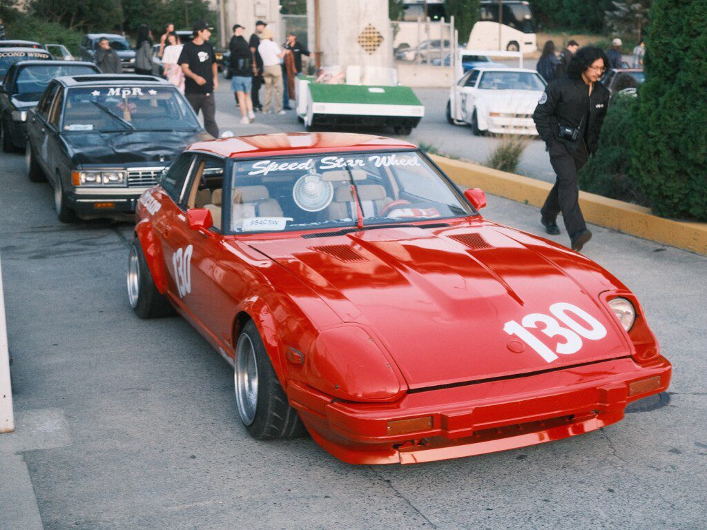Red classic race car number 130 at a car meet-up, surrounded by other vehicles and people in the background.