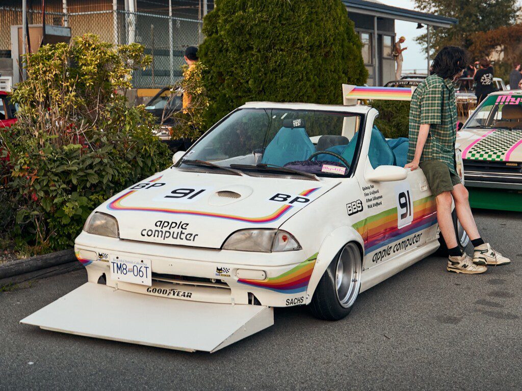 Retro-styled car with Apple Computer branding and rainbow stripes parked, person in checkered shirt leaning on it.