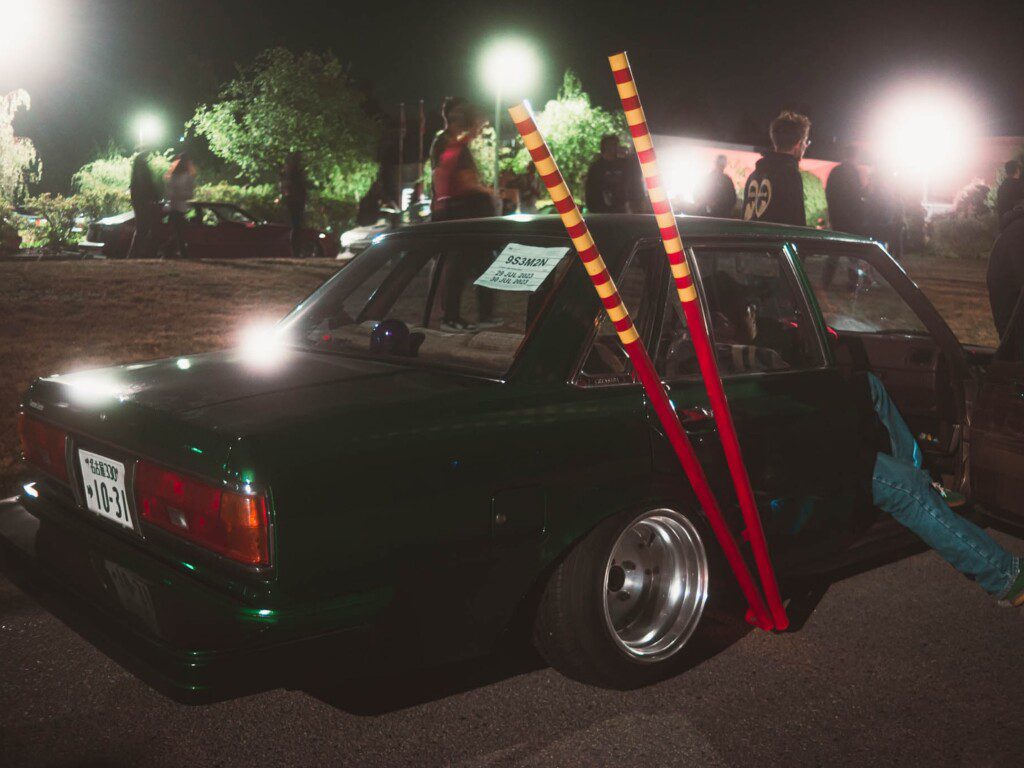 Modified car with red pipes at a night event, surrounded by people and lit by bright lights.