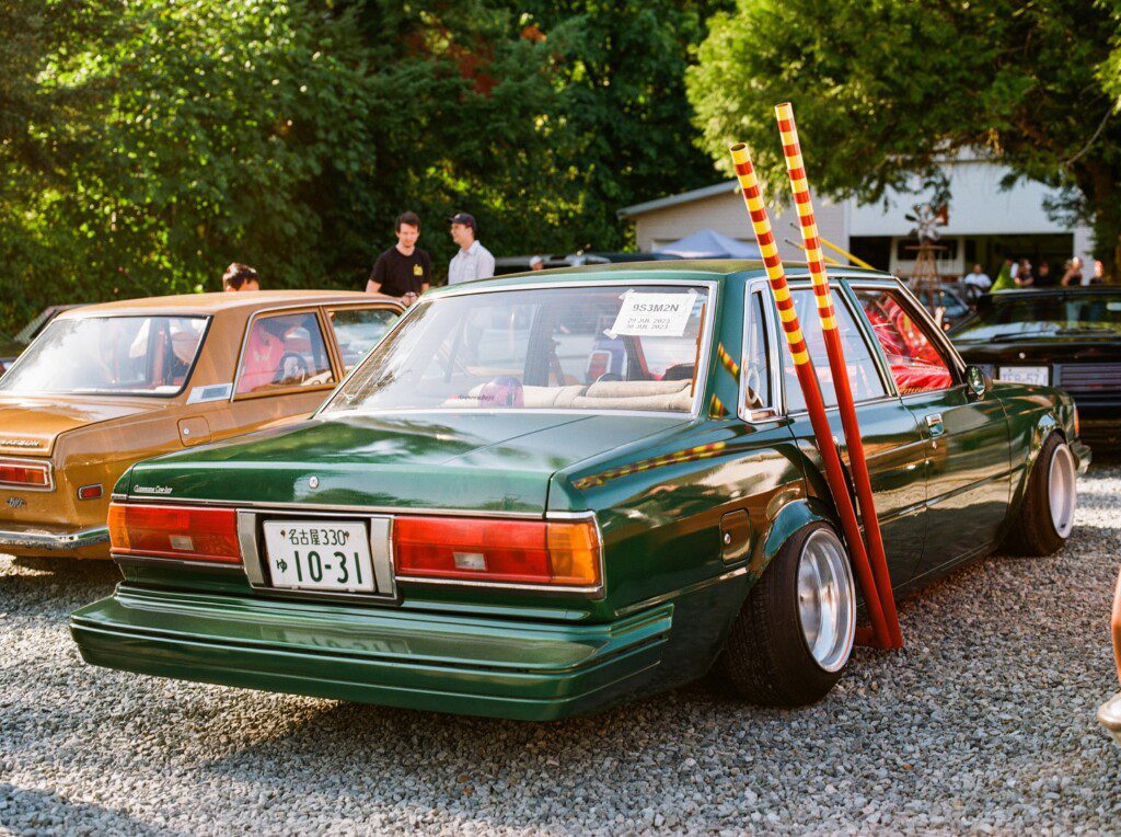 Vintage green car with bamboo poles, parked on gravel at an outdoor car meet, surrounded by people and trees.