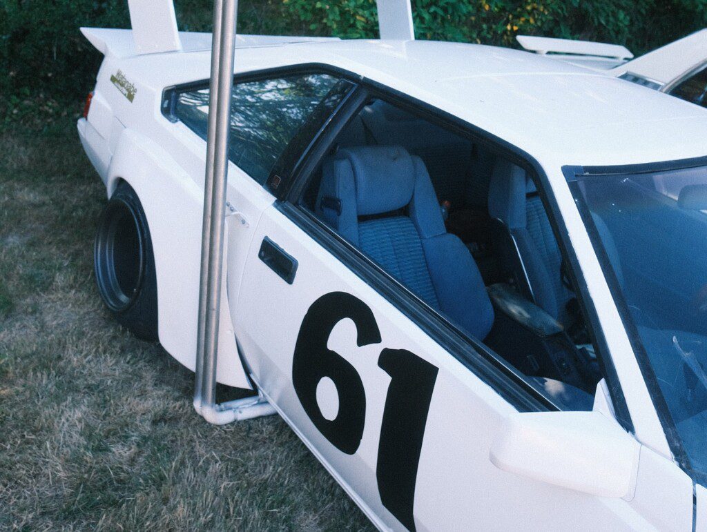 White vintage race car with number 61 on the door, parked on grass beside a green bush background.