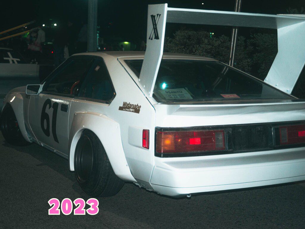 Vintage racing car with a large spoiler, number 61, parked at night. Text 2023 at the bottom.