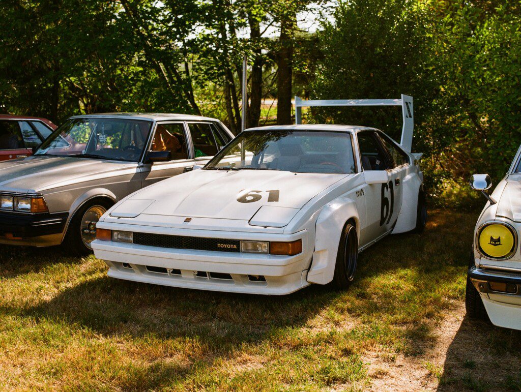 White Toyota race car with number 61 parked outdoors, surrounded by other vintage cars on grass.