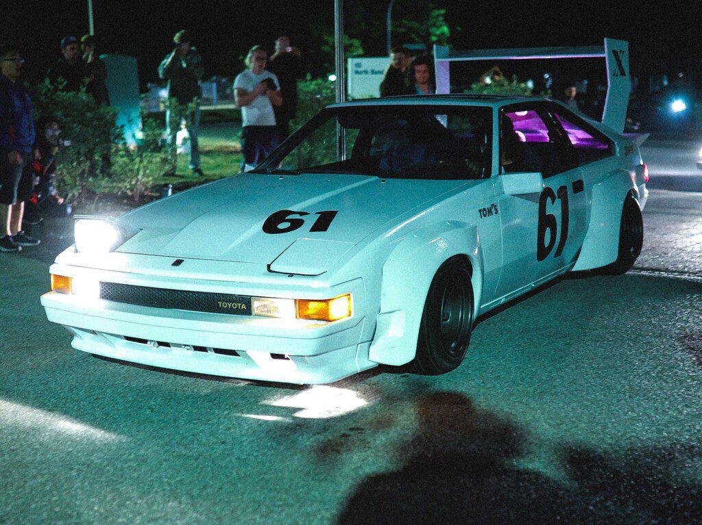 Vintage white Toyota race car with number 61 at night event, surrounded by people, showcasing classic style.