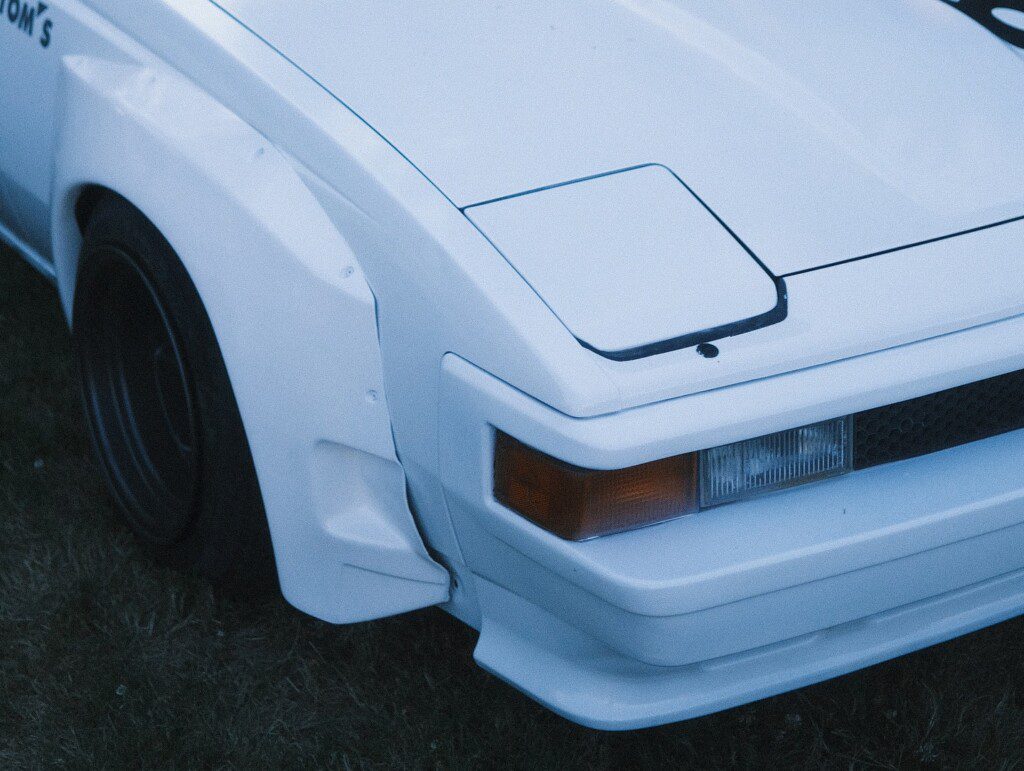 Close-up of vintage white sports car front with pop-up headlights and sleek design, parked on grass.