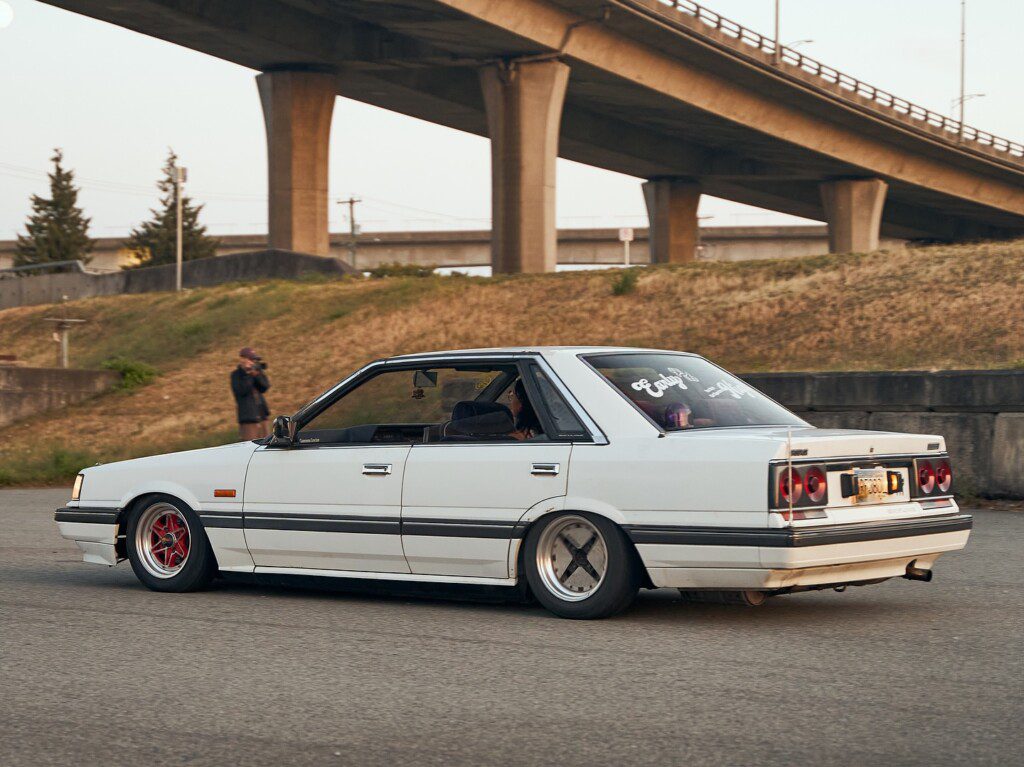 White vintage car with custom wheels driving under a bridge, accompanied by a photographer. Automotive photography scene.