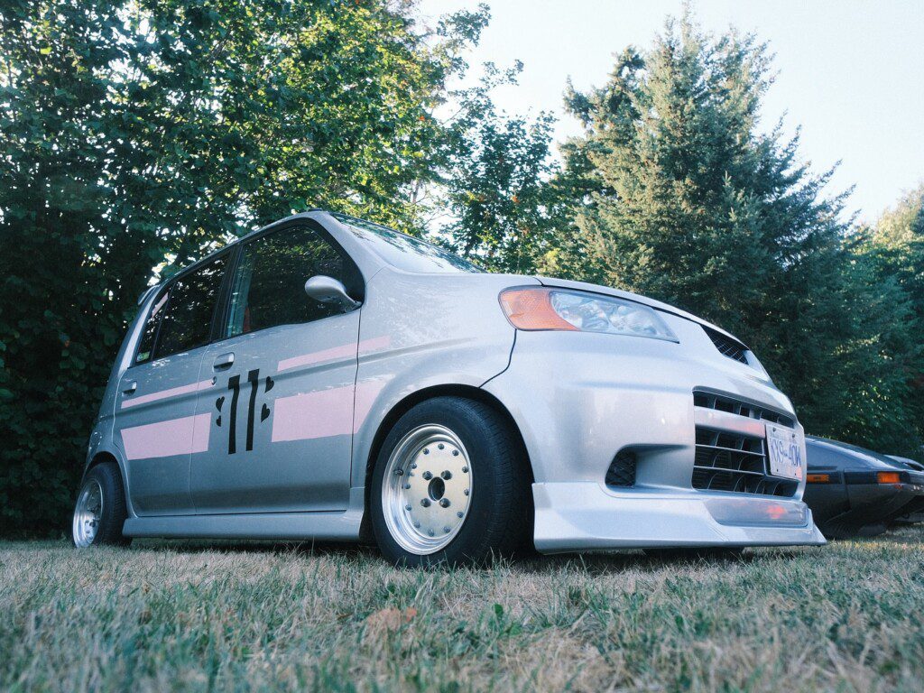Customized silver hatchback with pink decals parked on grass with trees in the background.
