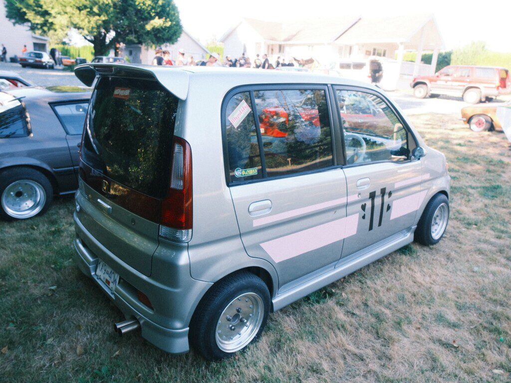 Silver compact car with sporty decals parked at a car show, surrounded by other vehicles and people.