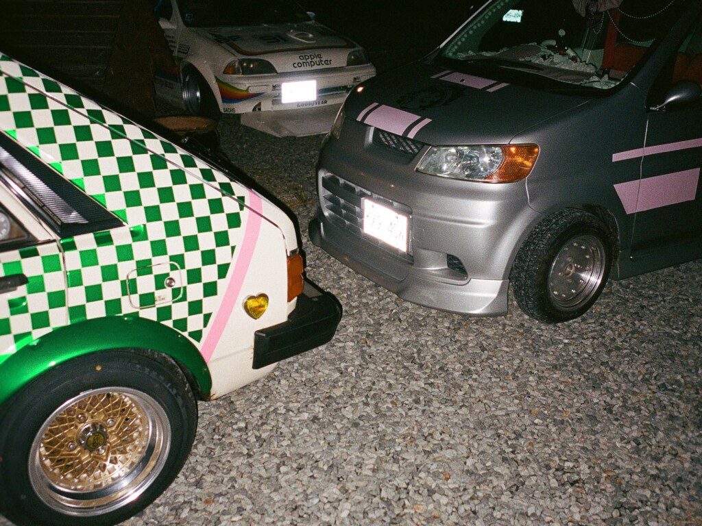Vintage cars with unique paint jobs parked on gravel at night, featuring checkered and striped designs.