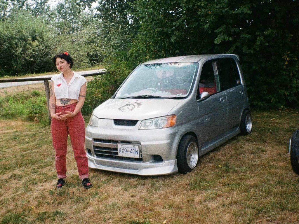 Person in vintage style near a modified compact car in a lush, green outdoor setting.