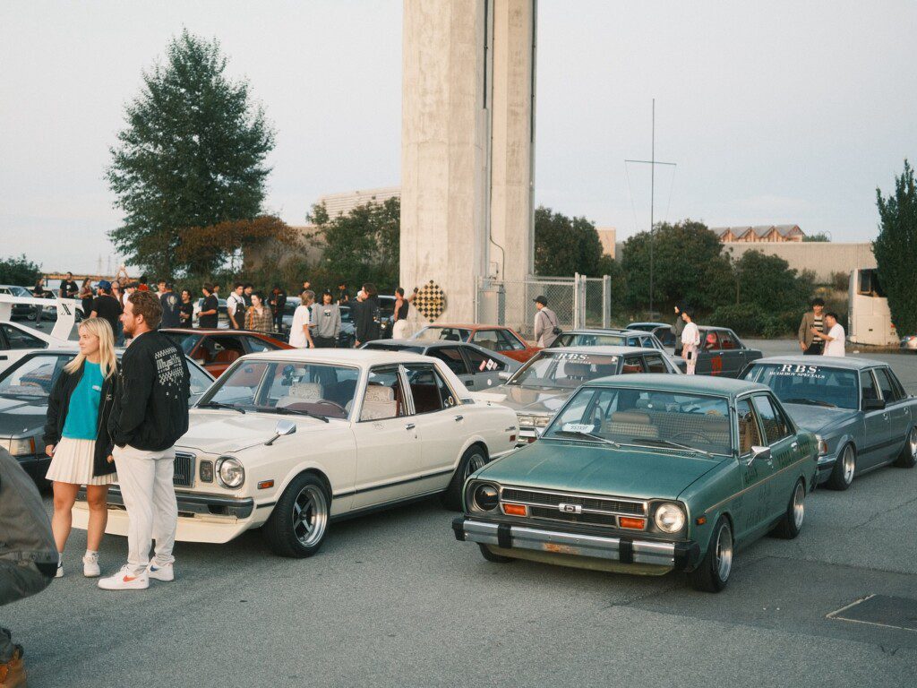Vintage car meet with classic sedans and a crowd socializing in a parking lot under a clear sky.