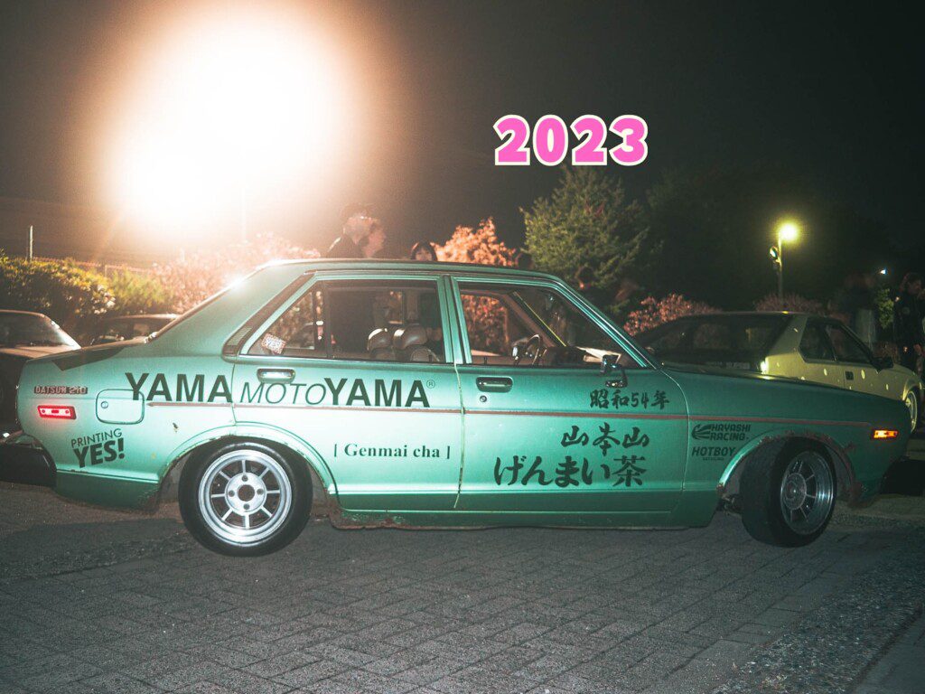 Vintage green Datsun car with modified decals, parked at a nighttime gathering in 2023.