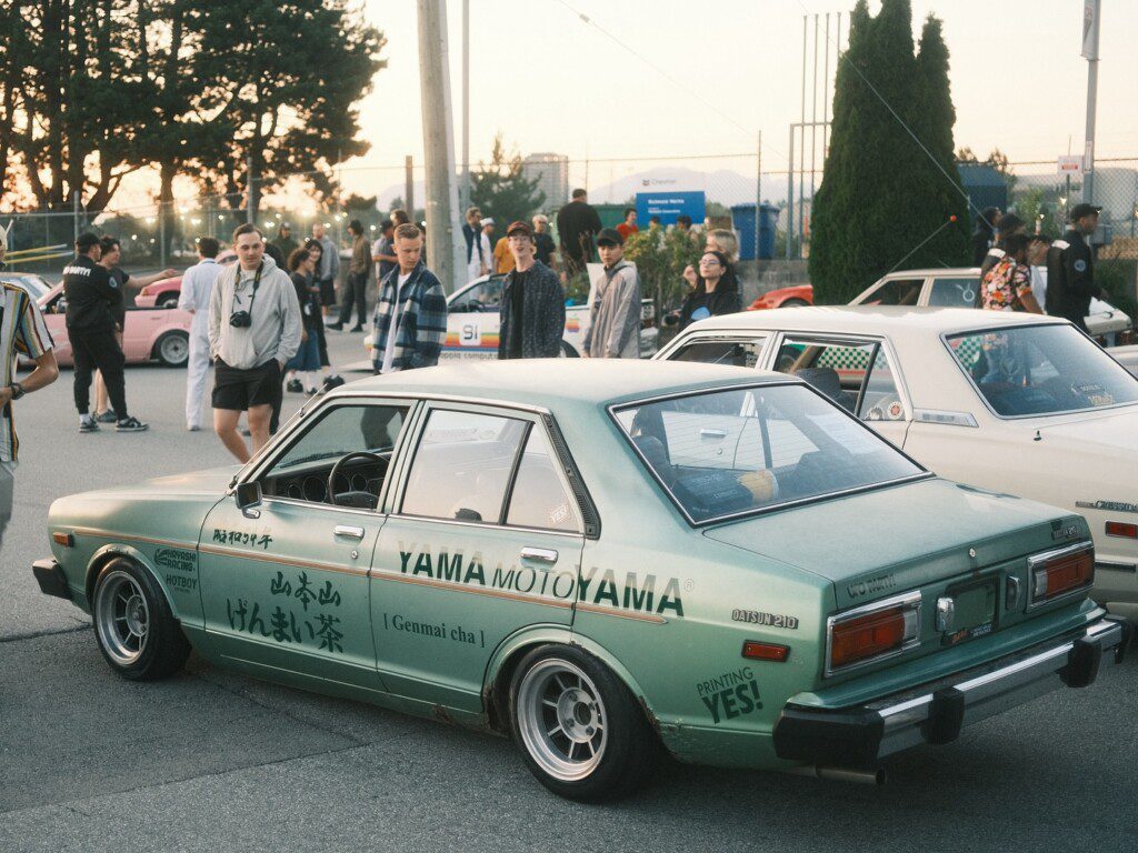 Vintage car meet with customized green Datsun 210, surrounded by people in casual attire at sunset.