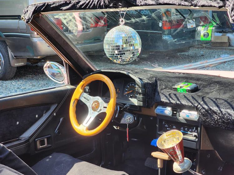Retro car interior with wood steering wheel, disco ball, fuzzy dashboard, and trophy cup on center console.