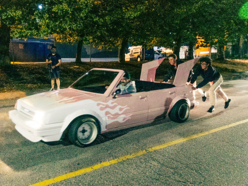 Group pushes a custom pink convertible with flames at night, while a person films the scene.