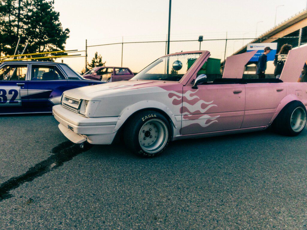 Pink convertible with flame decals at a car event, parked next to vintage cars under a sunset sky.