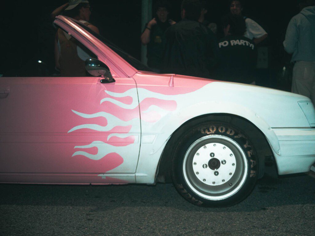 Vintage car with pink flame design and Goodyear tire on a night street, people gathered in the background.