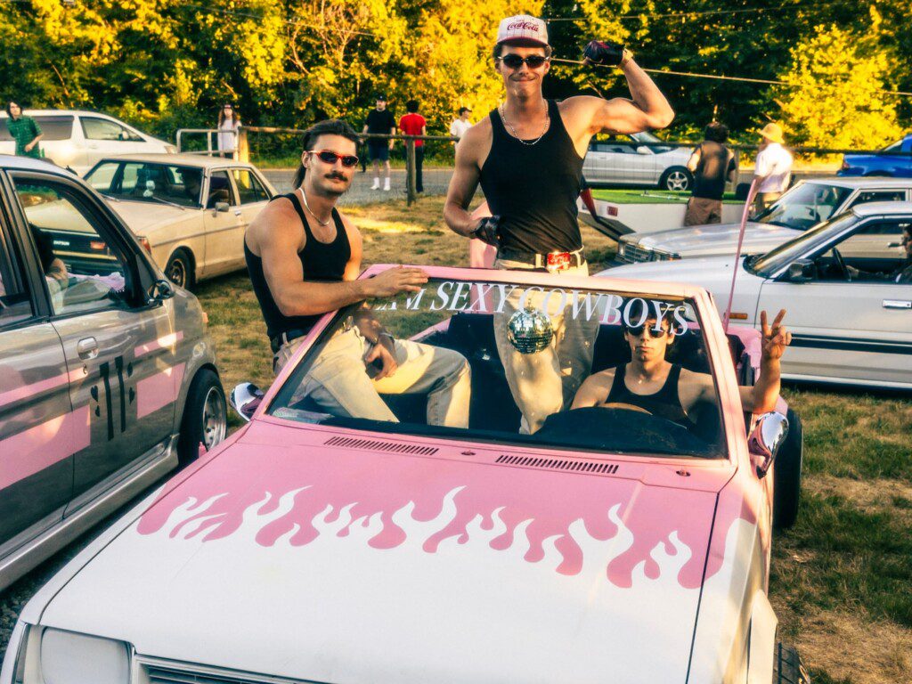 Group posing with pink car featuring flames and Sexy Cowboys decal at outdoor event.