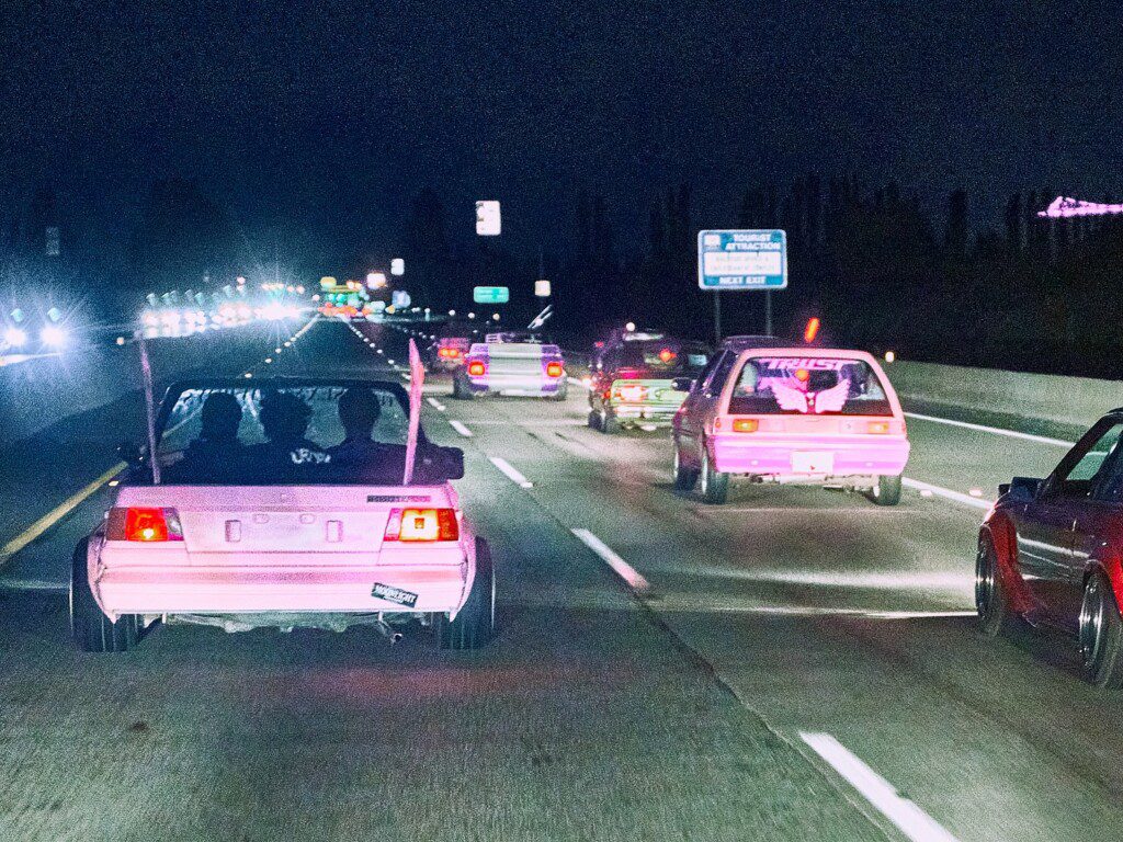 Modified cars cruising on a highway at night, showcasing vibrant pink lights and customization.