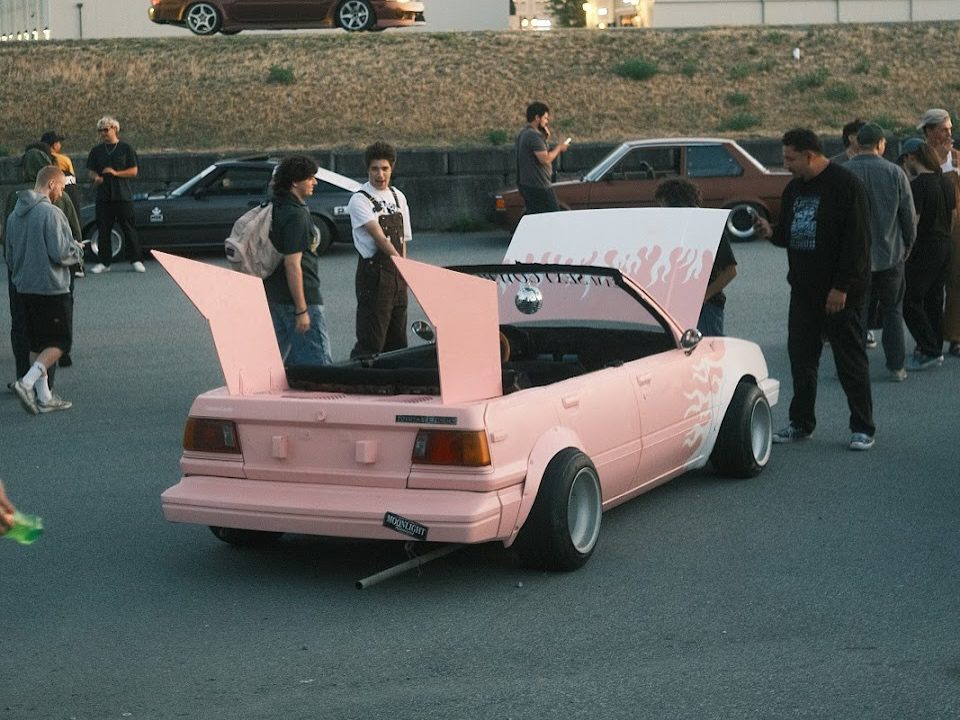 Pink modified car with raised trunk and hood at an outdoor car meet, surrounded by people.