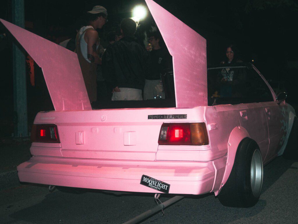 Pink Toyota Tercel with custom wings at night gathering, showcasing unique automotive modification.