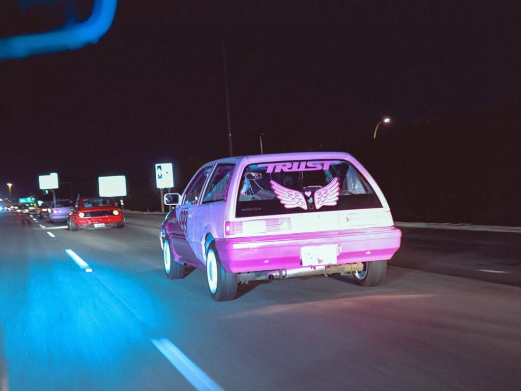 Retro pink car with wing decals and TRUST text cruising at night on a highway, followed by other vehicles.