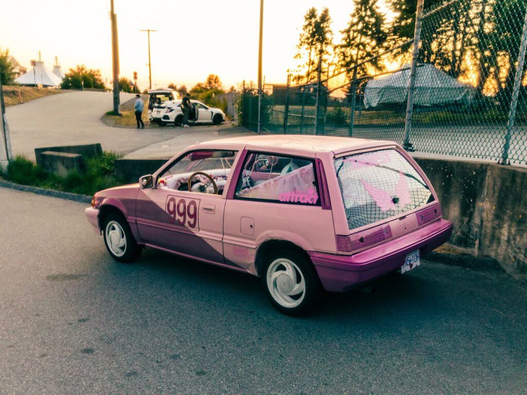 Pink hatchback with number 999 parked near a fence at sunset, with decals and unique design elements.