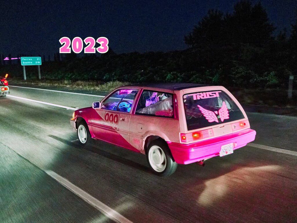 Pink retro car with wings decal cruising a highway at night, 2023 text above, city signs visible.