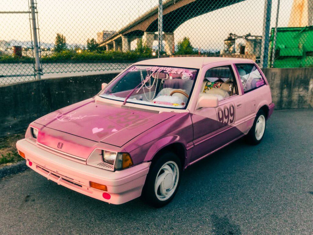 Pink vintage car adorned with hearts and stickers, parked near a chain-link fence under a bridge at sunset.