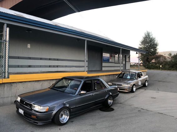 Two classic cars parked outside a warehouse under a bridge during sunset.